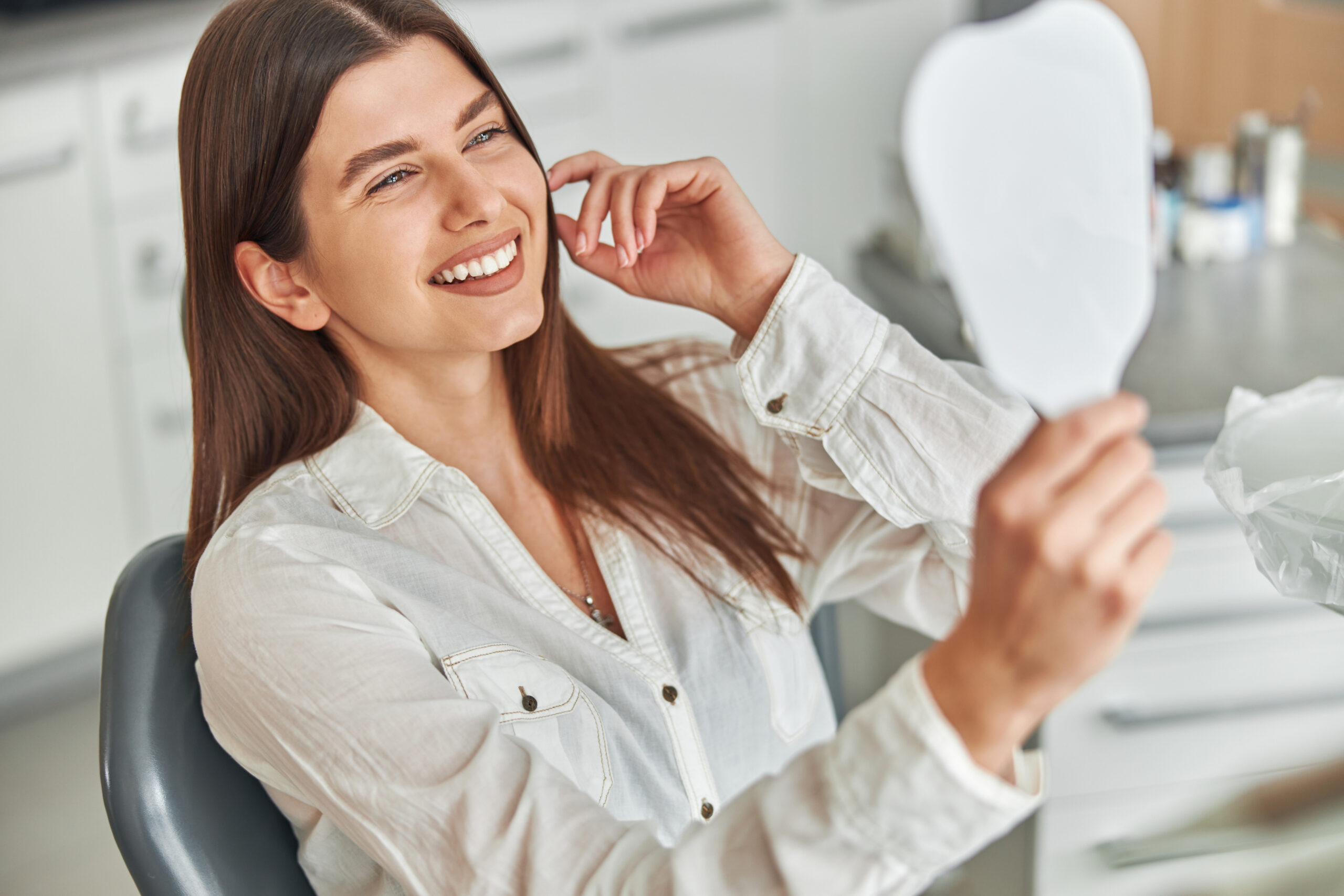 Happy young woman smiling checking out her perfect healthy teeth in the mirror, sitting in a dental chair at the dentist office